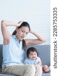 Small photo of Asian young stressed mother holding head with hands sitting with 7 months crying pitiful infant baby sitting on sofa at home. Single mom suffering from headache. Child care, Difficulties of motherhood