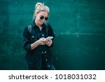 Smiling caucasian young woman sending message on smartphone standing near promotional copy space background, positive trendy hipster girl blogging in social network using mobile phone with internet