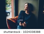 Thoughtful young woman dressed in black casual outfit looking out of window resting in comfortable coworking space. Pondering hipster girl with smartphone in hands thinking on future