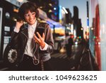 Cheerful pretty young woman in cool eyeglasses and trendy wear walking on metropolis street with night lights enjoying online playlist songs in earphones and reading sms with good news on smartphone