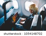 Attractive caucasian female passenger of airplane sitting in comfortable seat listening music in earphones while working at modern laptop computer with mock up area using wireless connection on board