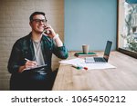 Cheerful male freelancer making telephone call share good news about project working in cafe interior,happy hipster guy having smartphone conversation while studying in good mood writing in planner