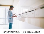 Small photo of Pensive hipster girl in casual wear using key for unlock mailbox in apartment hall, woman checking notification on mobile phone open metal letterbox for get correspondents from postal service