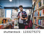 Handsome hipster guy in spectacles making money transaction for buying new bestseller via mobile phone, caucasian stylish man spending time in bookstore while browsing internet on smartphone