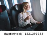 Young caucasian smiling female enjoying her comfortable flight while sitting in airplane cabin, listening to music in earphones and drinking water. Wifi internet access on board, passenger near window