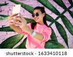 Small photo of Happy millennial content maker shooting influence video vlog near photo zone enjoying networking lifestyle, cheerful Asian hipster girl in sunglasses smiling at smartphone camera for selfie image
