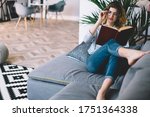 Full body woman with colored loose hair with glasses reading with interest book in comfortable pose on couch in living room at home