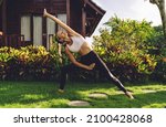 Small photo of Full body of fit female in sport outfit doing exalted crescent lunge yoga exercise in bright garden near small wooden house in warm summer day