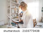 Small photo of Focused mature european female entrepreneur writing something in clipboard. Small business and entrepreneurship. Modern successful woman. Home art studio with pottery on shelves at sunny day