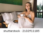 Small photo of Portrait of cheerful woman enjoying time for drinking tasty caffeine beverage, happy Caucaisan female in silk nightgown holding tea cup smiling at camera in hotel room for relaxing and resting