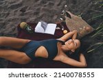 Young female in stylish swimwear enjoying recreation time at coastline beach - nap during leisure for dreaming, happy woman with closed eyes sleeping at seashore - pastime for resting in swimsuit