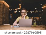 Smiling young female freelancer with TWS earbuds and eyeglasses look at camera with hand at chin while listening to music during break at table with laptop smartphone diary pen glass of juice in cafe