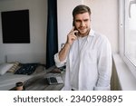 Small photo of Pensive young male entrepreneur looking away while standing at table with laptop throwaway coffee cup and talking on mobile phone in daylight near window