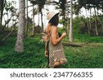 Small photo of Side view of young faceless female with handbag and straw hat standing in green park while enjoying lush green grass and tress against blurred forest during holiday