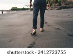 Back view of crop unrecognizable barefoot person walking along wet sandy beach against blurred background while enjoying vacation alone on seashore