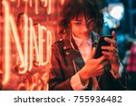 Young stylish female hipster standing near neon red light in night city and listening to music in headphones on modern smartphone. Woman watching media video on cellphone device