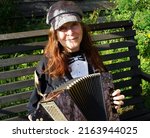 Small photo of Elderly woman with red hair and a cap sits outside and plays a diatonic button accordion