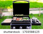 Small photo of An opened gas grill with vegetables, meat, and sausages in aluminum barbeque trays or dip pans. The background with green grass, bushes, a sidewalk are blurred. copy space on the black grill cover.