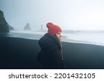 A woman in a black jacket and winter hat is standing on a black beach in Iceland. Hazy rocks and waves in the background. Misty rainy day.
