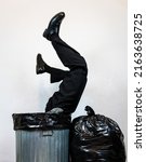 Small photo of Businessman in Suit Stuck Upside Down in Metal Trash Can Next to Garbage Bag Pile. Concept of Over a Barrel. Thrown Away by Capitalism and Greed.