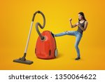 Small photo of Young woman in gray top and blue jeans, fists clenched and raised, kicking big red vacuum cleaner with right leg on yellow background. Tired of domestic chores. Thorough cleaning. Free trial period.