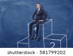 Small photo of Businessman is sitting on the first place of a podium on blue blackboard background. Succes. Winning in competition. Best solutions