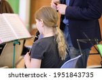 Small photo of A little girl with long blonde hair pulled back in a ponytail in the classroom in music class looks at the notes and plays the clarinet to the teacher standing next to her. School education concept