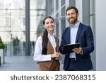 Small photo of Two happy male and female business colleagues in suits standing outside near office, discussing important work issues, comparing papers, preparing for presentation, looking at camera and smiling.