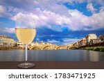 Glass of white wine with view of bridge in Florence during sunset - Ponte alla Carraia,  five-arched bridge over Arno River in the Tuscany region of Italy. Beautiful blue with pink sky with clouds. 