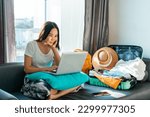 Asian woman prepare things into baggage for vacation trip at home. Happy female using computer to planning travel trip and booking tickets or hotel room on web. Travel, summer and holiday concept.
