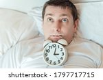 Insomnia. Man lies in bed with open eyes and alarm clock in mouth trying to fall asleep at night. Counting sheep awake. Stress thoughts in bedroom with white linen. Sleep disorder.