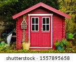 A light red small shet, gardenhouse, with some garden tools around it
