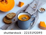 Mug of tasty Pumpkin soup with seeds and crackers. healthy orange Pumpkin cream soup in a grey cup with pieces of pumpkin on a white kitchen table. Vegetarian autumn soup. copy space.
