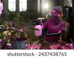 Small photo of Unrecognizable woman with arm prosthesis wearing protective kerchief mask and goggles spraying plants with water in post-apocalyptic farm