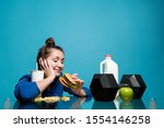 Small photo of the girl with undisguised pleasure looks at the burger in her hand while in front of her lies a sports equipment and healthy food, copy space