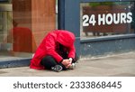 Small photo of A young person sat on a street pavement curled up in a red hooded jacket and head bent right down. The shop window behind advertises 24 hours which would feel like an eternity when you are in crisis .