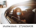 Woman wearing a medical sterile mask in taxi car on a backseat looking out of window checking her cell phone. Girl passenger waiting in a traffic jam during coronavirus quarantine. Healthcare concept