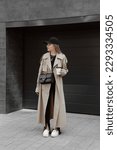 Small photo of beautiful blonde girl dressed in beige trench coat, black longsleeve, flared pants with slits, white loafers, cap, shoulder bag, accessories, stylish fashion outfit, full length lifestyle model