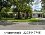 Facade of a modern and luxurious colonial-style house in the city of Coral Gables, Miami-Dade, USA, with large tropical vegetation around it, open garage, main entrance, sidewalk, short grass, privet 
