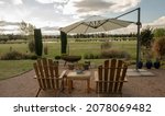 Small photo of Relaxation. View of the beautiful garden with wooden exterior furniture, garden chairs and table, and big sunshade umbrella at sunset.