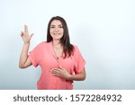 Small photo of Cheerful caucasian young woman keeping hand on up, loke gun, holding other on stomach isolated on white background in studio in pink shirt . People sincere emotions, lifestyle concept.