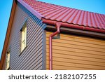 Small photo of Corner of the house with metal tile roof and rain gutter. Red gutter on the roof top of house. Rooftop water collection system. Metallic Guttering System, Guttering and Drainage Pipe Exterior