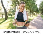 Small photo of Energize your morning. Cheerful fit woman in sportswear refreshing, drinking water and using her smartphone after jogging in a green park on a sunny day