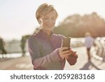Small photo of Results speak for themselves. Portrait of joyful active mature woman in sportswear using her smartphone, checking result after running outdoors on a sunny day