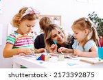 Small photo of Little girls and smiling educator studying geometric figures, playing and folding cubes on desk sitting on floor in playroom. Interesting lesson for kindergartners developing logic and intelligence.