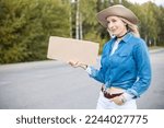 Modern, grimacing, sarcastic blond woman in hat hitchhiking with blank carton board and waiting for car stop. Travelling tourist. Hitchhiker risks and dangerous trip. Adventure autostop lifestyle