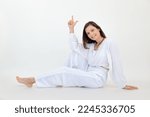 Small photo of Portrait of smiling beauteous young woman with short dark hair wearing white loose blouse, trousers, sitting on floor, raising index finger up on white background. Sport, yoga, advertisement. Studio.