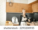 Small photo of Portrait of young smiling beauteous woman wearing dirty white apron with dots, grey sweatshirt, standing in kitchen at home, looking happily. Unrecognizable man holding glass bowl. Cooking, baking.