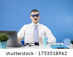 Small photo of Young handsome Caucasian cheerful man dressed in a classic shirt with a blue tie looks in surprise at the camera and opens his mouth wide. Ludicrous funny blue sunglasses. Stylish office interior.