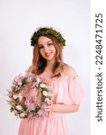 Small photo of A girl in a pink dress and a wreath on her head is holding Easter wreath with eggs on a white background. Smiling young woman holding Easter wreath with eggs on a white background.